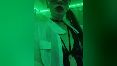 Video of a woman with light skin and glasses, wearing a white shirt and black lace lingerie, tongue out, in a dimly lit room with green LED lights.