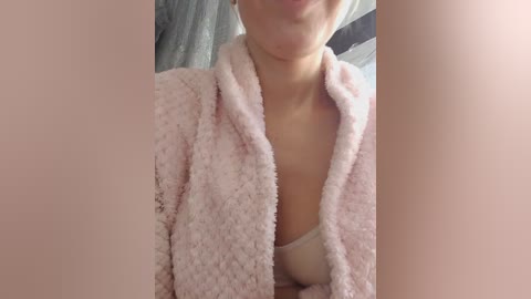 Video of a light-skinned woman with a slight smile, wearing a pink, textured robe with a white bra visible underneath, in a softly lit, indistinct indoor setting.