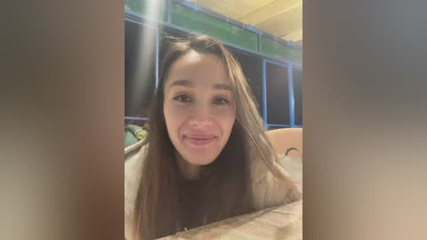 A video of a smiling young woman with long brown hair, wearing a beige sweater, indoors with a green curtain and a wooden table in the background.