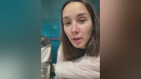 Video of a young woman with shoulder-length dark hair, wearing a beige quilted jacket, sitting at a table with a glass of water, in a dimly lit room with a teal wall.