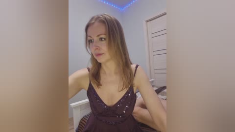 Video of a fair-skinned, slim woman with shoulder-length brown hair, wearing a dark purple sequin dress, sitting on a bed in a bedroom with blue LED lights and a white door in the background.