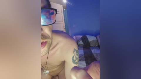 A video of a shirtless man with a tattoo on his shoulder, wearing VR glasses, lying on a bed with a striped blanket. The background shows a blue wall and a partially visible door.