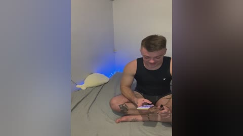 Video of a muscular, fair-skinned man with short brown hair, wearing a black sleeveless shirt and gray shorts, sitting cross-legged on a gray bed, playing with a stuffed animal, with blue LED lights in the background.