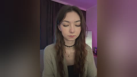 Video of a young woman with long brown hair, wearing a beige cardigan over a black top, a black choker, and closed eyes, set against a dimly lit room with purple and pink lighting.