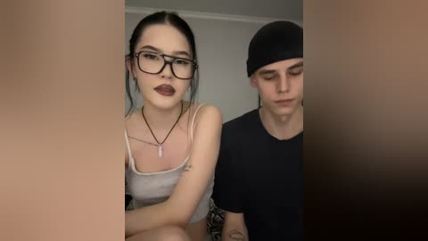 Video of a pale-skinned, black-haired woman with glasses and dark lipstick, wearing a white tank top, and a tattooed man in a black beanie and t-shirt.