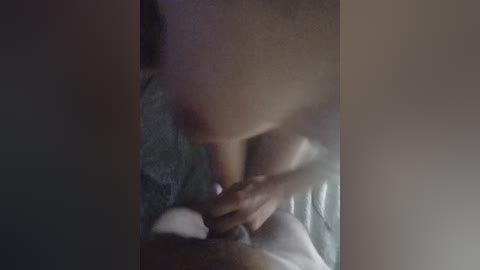 A blurred video of a nude, light-skinned woman, possibly Latina, with medium-sized breasts, crouching over a man's erect penis, partially obscured by her hand. The background is dimly lit, with a textured surface visible.