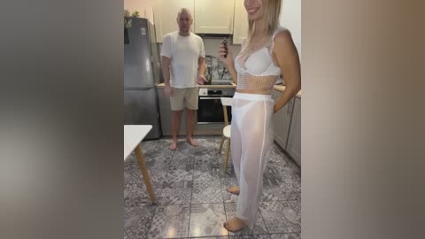 A video of a blonde woman in a sheer white lace bodysuit and matching pants, standing in a modern kitchen, holding a phone, while an older man stands behind her.