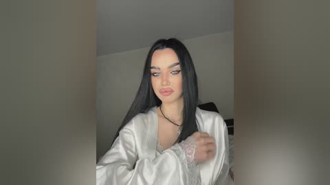 Video of a young woman with long black hair, wearing a white satin robe, standing indoors. She has a pale complexion, green eyes, and a confident expression. The background is plain and dimly lit.
