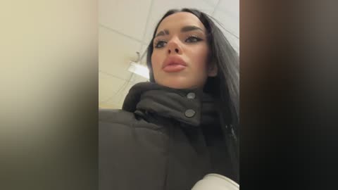A video of a woman with long, straight black hair, wearing a black puffer jacket, and looking directly at the camera with a confident expression. The background is a plain, beige-colored wall.