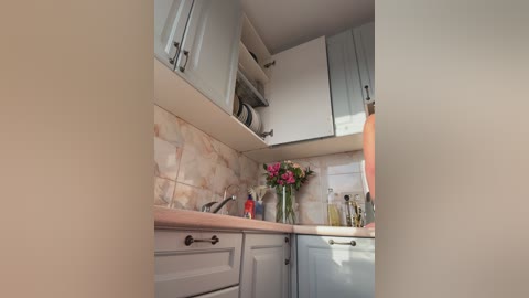 Video of a small, modern kitchen with light gray cabinets, a marble backsplash, and a stainless steel sink. A bouquet of flowers sits on the counter next to a red bottle, and a white towel hangs on a hook.