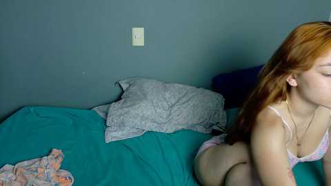 Video of a fair-skinned woman with long red hair, wearing a white bra and panties, lying on a teal bed with gray pillows and a floral-patterned cloth.