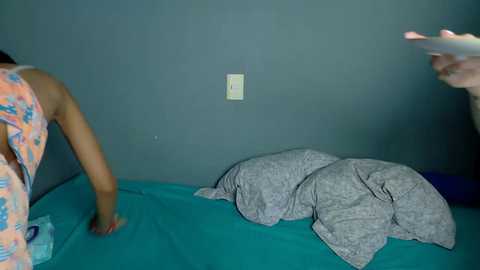 Video of a person in a colorful floral nightgown standing by a teal bed with gray pillows and a light switch on a teal wall.