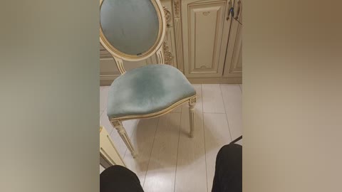 Video of a vintage, pale blue upholstered chair with ornate, cream-colored wooden frame and intricate carvings, set against a beige wall with decorative paneling and a white tiled floor.