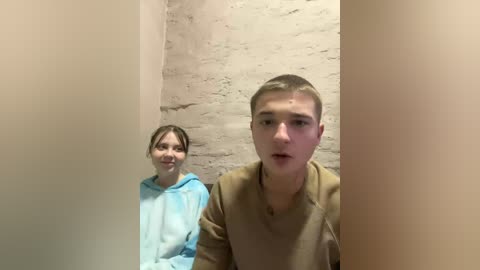 A video of a young boy and girl in a dimly lit, narrow, brick-walled corridor. The girl, with light skin and brown hair, wears a light blue hoodie. The boy, with short blond hair, has a neutral expression.