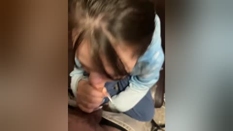Video of a young girl with dark hair and light blue jacket kneeling on a wooden floor, looking down with hands clasped in a prayer-like pose. Background shows a blurred, dark room.