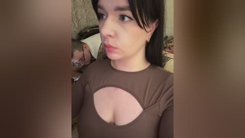 A video shows a young woman with fair skin, dark hair, and a brown top with a cutout revealing cleavage, seated next to an older man with glasses. The background is a beige wall with a floral pattern.