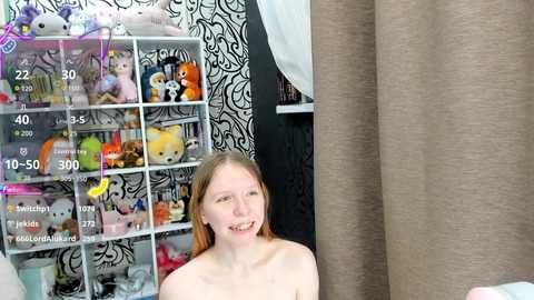 Video of a smiling, fair-skinned, topless woman with long, light brown hair, sitting in a room with a black and white patterned wall, a toy-filled shelf, and beige curtains.