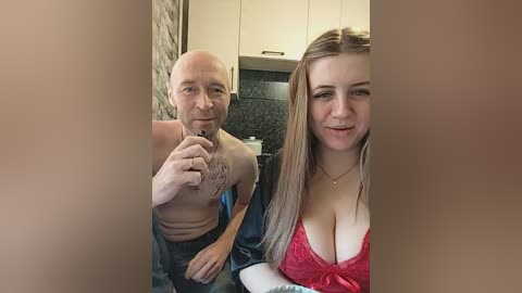 Video of a bald, shirtless, muscular man and a blonde woman in a red lace bra, sitting on a bed in a modern kitchen.