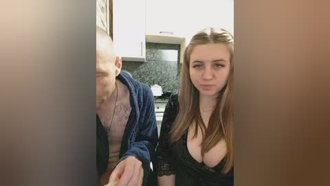 A video of a young woman with long brown hair and a light complexion, wearing a revealing black dress, standing beside a bald man in a blue robe, in a modern kitchen with white cabinets and a dark countertop.