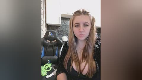 A video of a young woman with long, light brown hair, wearing a low-cut black top, sitting in a gaming chair with a headset and controller nearby in a modern kitchen.