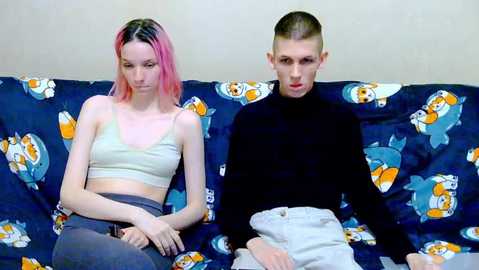 Video of a pale-skinned woman with pink hair, wearing a light green crop top and grey pants, and a man with short hair in a black turtleneck, sitting side by side on a blue couch with cartoon fish pattern.