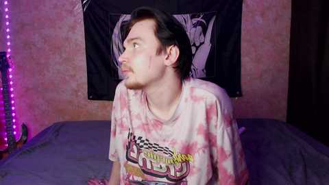 Video of a young man with pale skin, short dark hair, and a beard, wearing a tie-dye T-shirt, sitting on a bed in a dimly lit room with pinkish-purple LED lights, black tapestry with a white anime character, and a guitar on the left.
