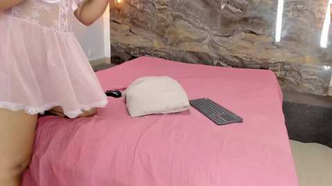 A video of a woman in a light pink, lacy babydoll dress, sitting on a pink bedspread. A beige pillow and a black remote control are on the bed. Background features a rustic stone wall.