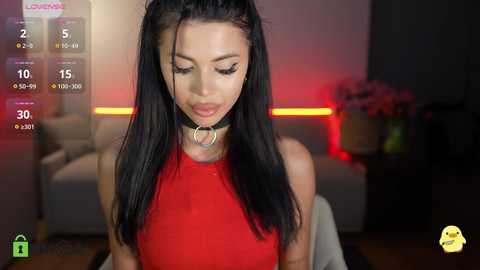 A video of a young woman with long black hair, wearing a red dress and black choker, smiling. Background shows a dimly lit living room with red lighting, and a blurred sofa.