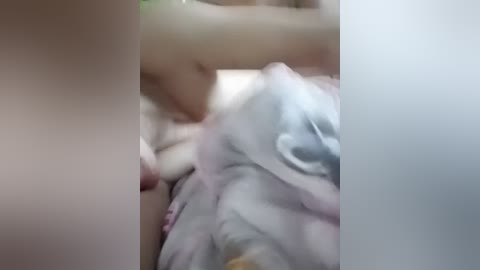 A blurred video of a woman lying on a bed, partially covered by a light-colored blanket, with a pale complexion and light hair.