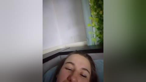 A video of a young woman with light skin and brown hair, eyes closed, lying on a bed with a light blue pillow. The background features a green plant and a white curtain, creating a serene, indoor bedroom setting.