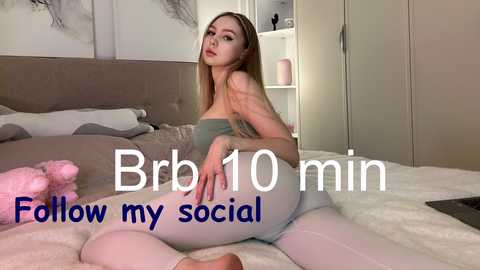 Video of a blonde woman with fair skin, wearing a strapless green top and tight white leggings, sitting on a bed with a grey headboard. Text overlays read \"Bra 10 min, show my social.\