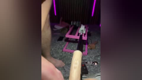 A video of a cluttered, dimly lit room with a pink, rectangular desk holding a black guitar. The floor is covered in scattered items, including a pair of shoes and cables, under dim purple lights.