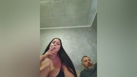 Video of a young woman with long black hair, large breasts, and pale skin, sucking on her finger in a dimly lit, tiled bathroom. A bearded man watches from the corner.