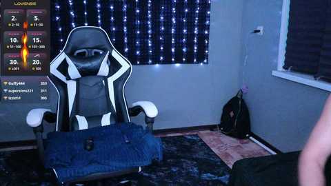 Video of a gaming room with a black-and-white gaming chair, a blue backpack, and a blue suitcase on the floor, lit by a string of white fairy lights on the wall.