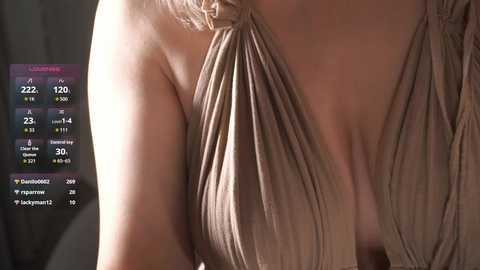 Video of a woman's bare chest in a beige, pleated, V-neck dress, with a digital screen displaying heart rate, respiratory rate, and blood pressure measurements in the background.