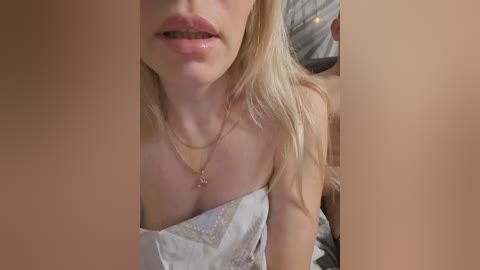 Video of a blonde woman with fair skin, wearing a light gray, lacy bra, lying on a bed with a white sheet. Her lips are slightly parted, revealing a hint of her teeth.