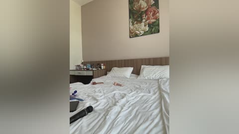 A video of a minimalist, neatly made bed with white linens and a brown headboard, positioned against a beige wall adorned with a large floral painting. A black hairdryer and a smartphone are casually placed on the bed.