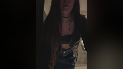 Video of a young woman with long, dark hair, wearing a black choker, black bra, and high-waisted leather pants, in a dimly lit, shadowy room.
