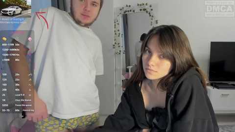 A video of a young man in a white t-shirt and green pajama bottoms standing behind a woman in a black robe, both in a modern, brightly lit room with a TV.