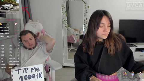 Video of a woman in a black robe sitting, holding a sign saying \"Tip me 40k,\" and a man in a white shirt and hat with a \"B\" on it, both in a modern, bright room.