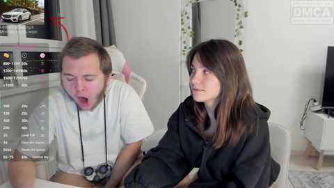 Video of a man with headphones on, looking shocked, and a woman in a black hoodie watching him, both in a modern living room with a TV and a mirror.