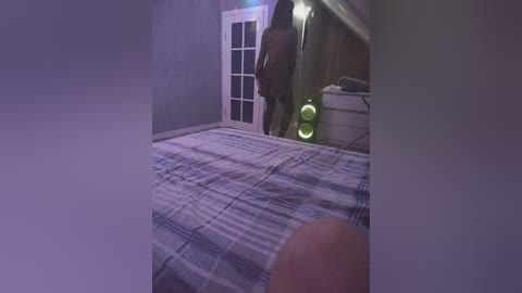 Video of a dimly lit bedroom with a large, plaid-patterned bedspread in shades of purple and white, a wooden wardrobe, and a small green light source.