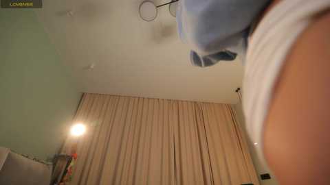 Video of a woman in a white tank top, with beige curtains, a green wall, and a lamp on the left.