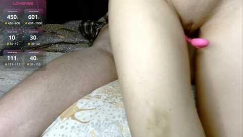 Video of a nude woman lying on her back on a bed, using a pink vibrator on her vulva, while a man's legs are visible in the background.