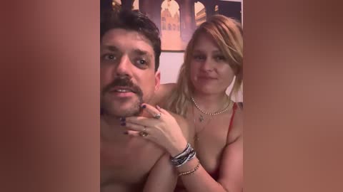 Video of a topless, bearded man with a mustache, and a blonde woman with long hair, wearing a necklace and bracelets, both smiling. Background shows framed artwork.