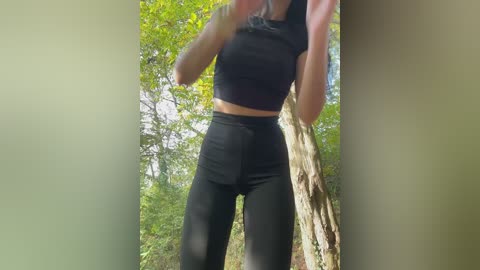 Video of a fit woman with light skin, wearing a black crop top and high-waisted black leggings, posing outdoors in a lush, green forest.