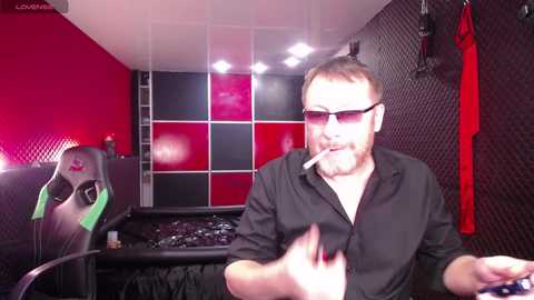 Video of a bearded man in glasses, wearing a black shirt and smoking, seated in a gaming chair in a dimly lit room with red and black walls, gaming equipment in the background.