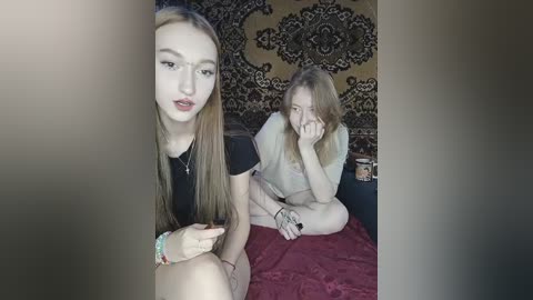 Video of two young women: one fair-skinned with long straight hair, wearing a black top, and another with light brown hair, holding her face, sitting on a red blanket.