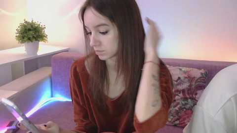 Video of a young woman with long brown hair, wearing a velvet burgundy top, sitting on a pastel sofa, holding a laptop, with a plant and colorful LED lights in the background.