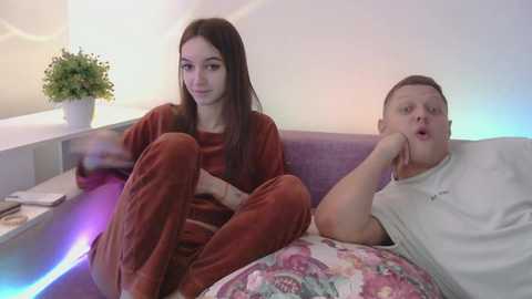 Video of a young woman with long brown hair and a young man with short brown hair, both wearing cozy red pajamas, sitting on a purple couch in a modern, brightly lit living room.